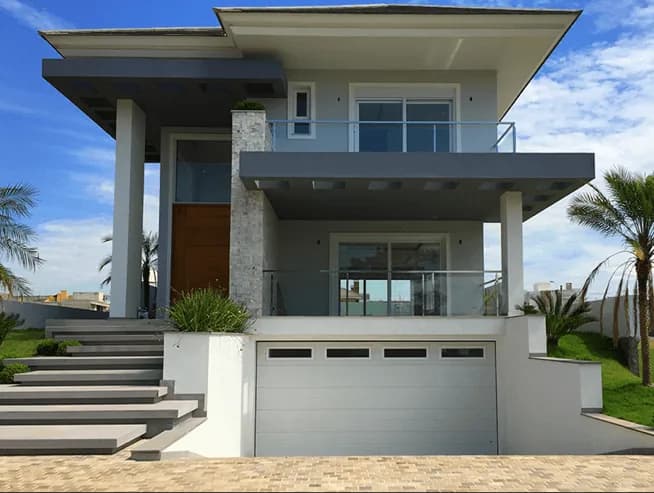 Casa de dois pavimentos com portão seccional branco na garagem, varandas com guarda-corpo e céu azul