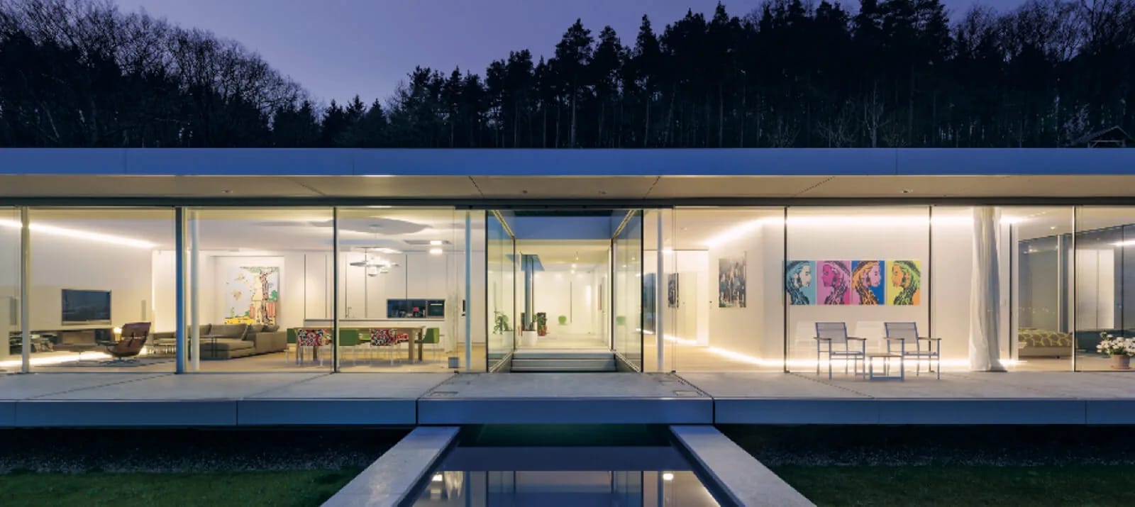 Casa térrea com fachada toda envidraçada, iluminada à noite, com espelho d'água em frente e mata ao fundo